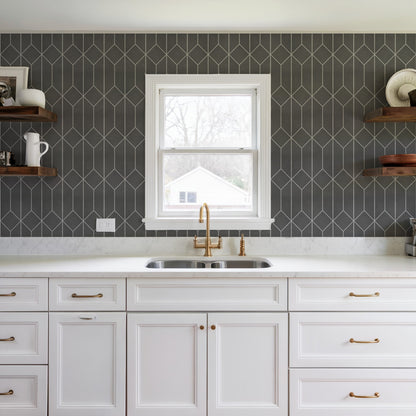 White kitchen cabinets with brass handles, marble countertop, dark geometric tile backsplash, double sink under a window, open shelves.