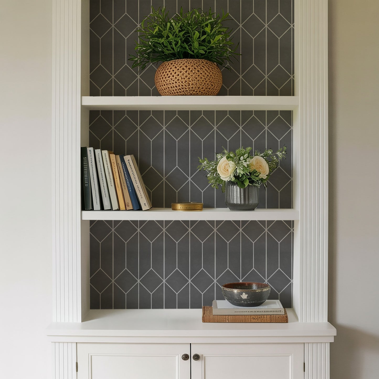 White built-in shelf with dark geometric wallpaper; top wicker pot, middle books with cream-rose vase, bottom bowl on books.