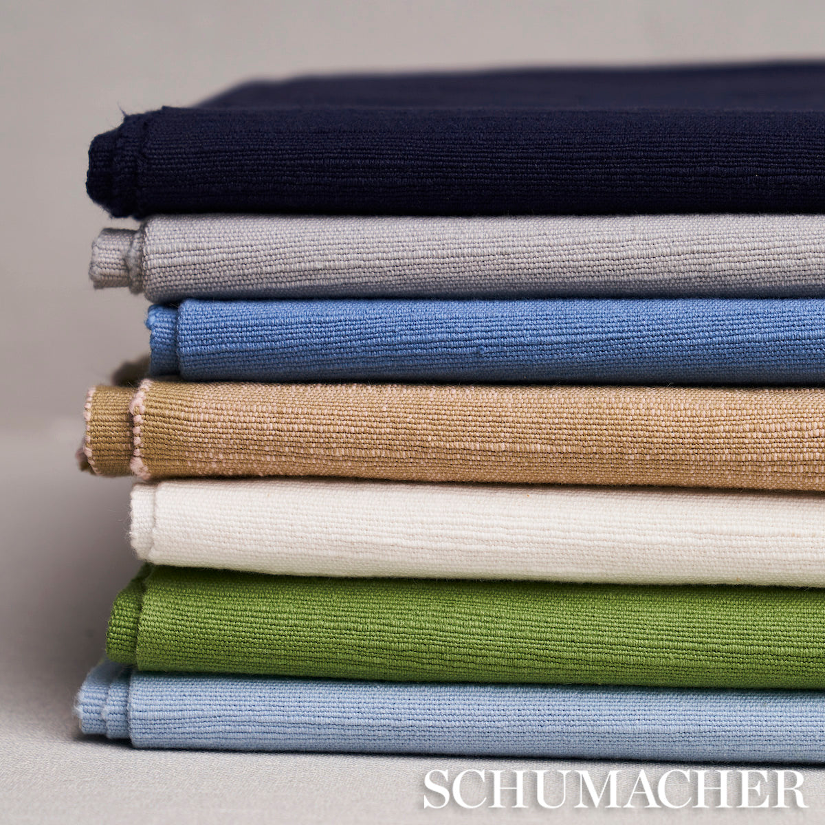Stack of folded woven fabrics in navy, gray, light blue, beige, white, sage green, and sky blue.
