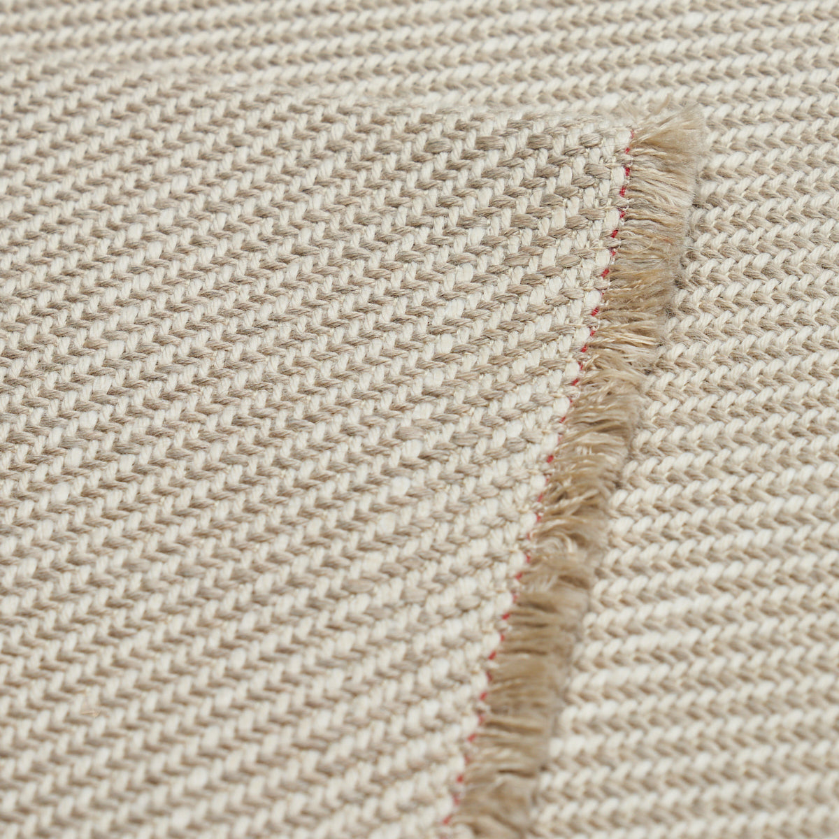 Natural beige woven rug with fringed edge and pink-red stitching along the seam; dense textured weave.
