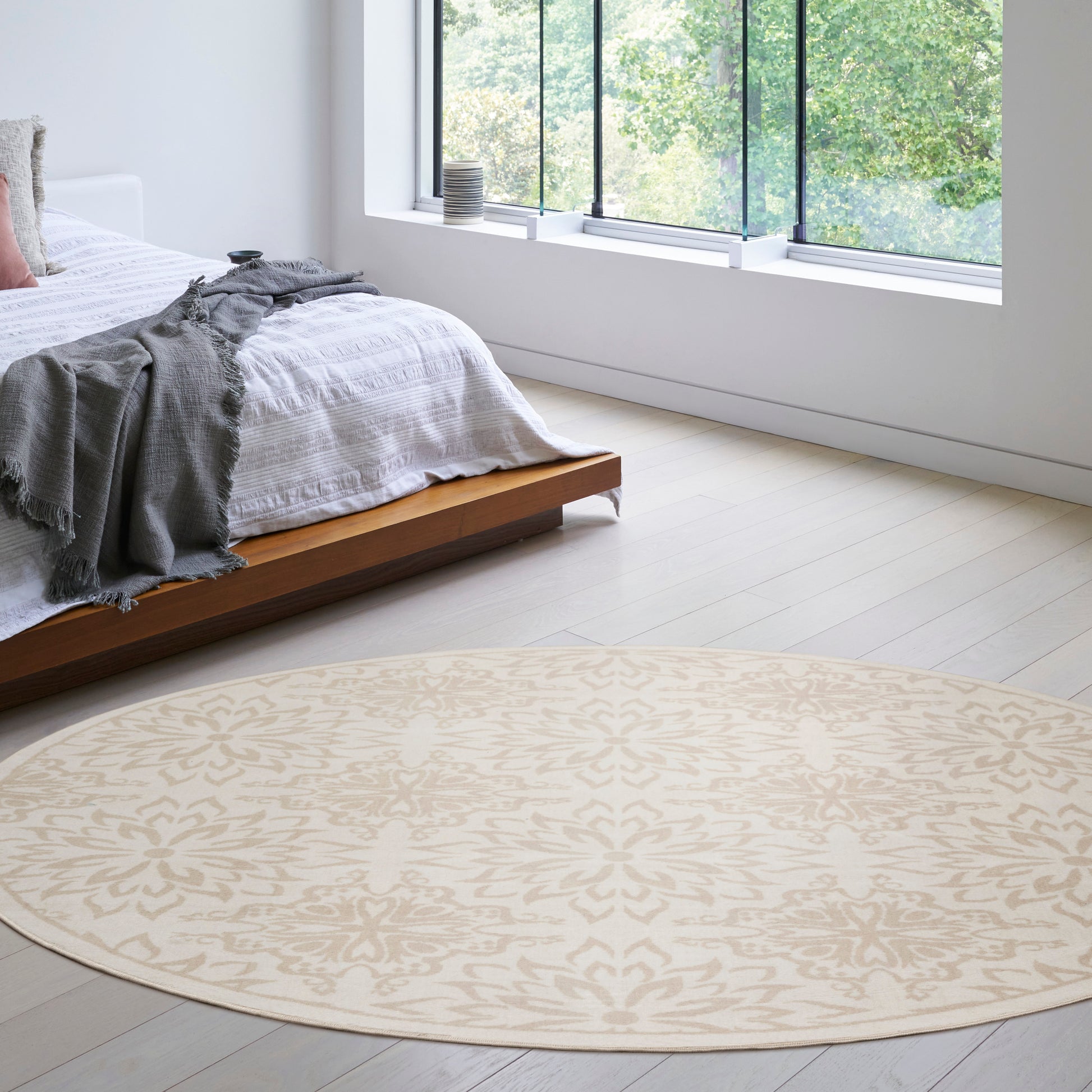 Round beige patterned area rug on light wood floor in bright modern bedroom with low wooden bed and gray throw.