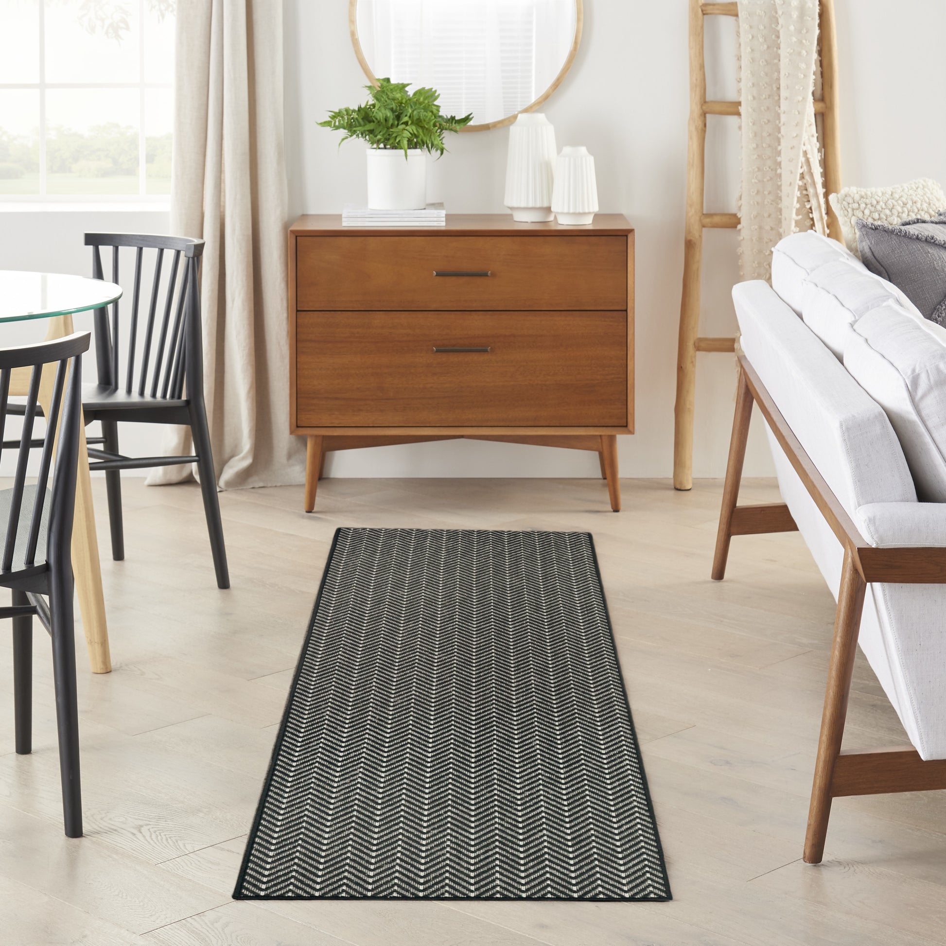 Dark chevron-patterned runner rug on light wood floor before a mid-century oak dresser with two drawers and a potted plant.