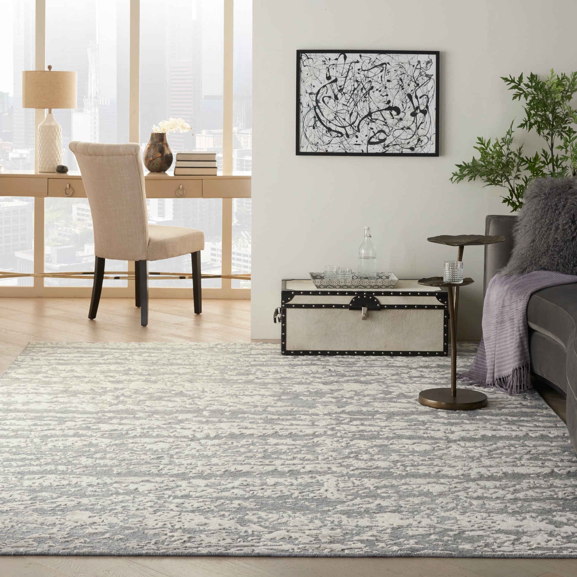Gray and white abstract area rug in a modern living room with beige upholstered chair, decorative trunk, and greenery.