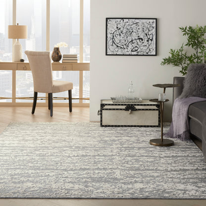 Gray and white abstract area rug in a modern living room with beige upholstered chair, decorative trunk, and greenery.