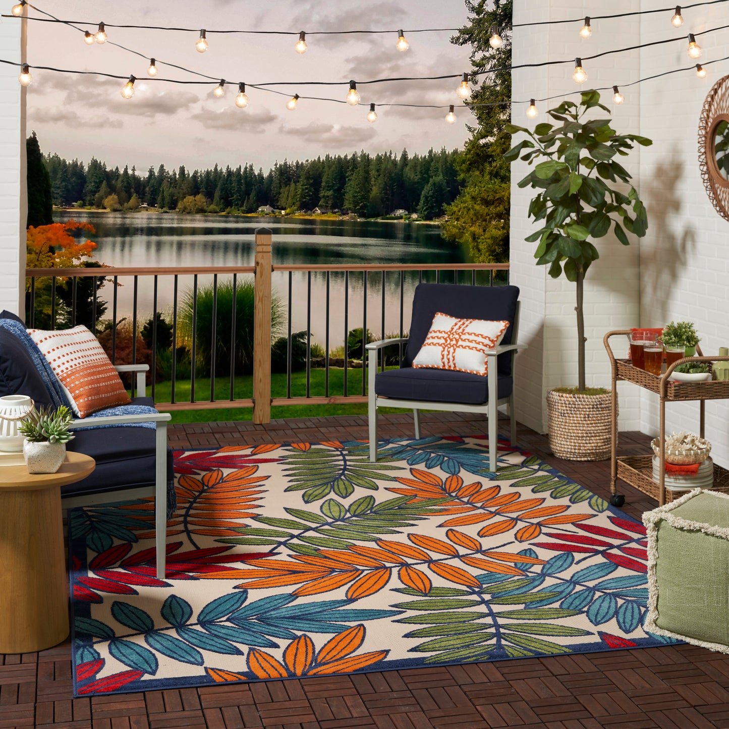 Outdoor patio with string lights, railing overlooking a lake, navy chairs, colorful leaf-pattern rug, and potted greenery.