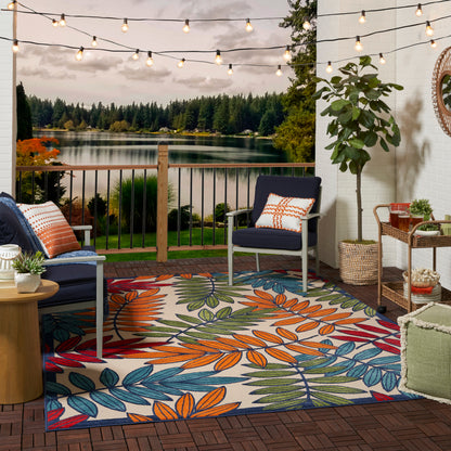 Outdoor patio with string lights, railing overlooking a lake, navy chairs, colorful leaf-pattern rug, and potted greenery.