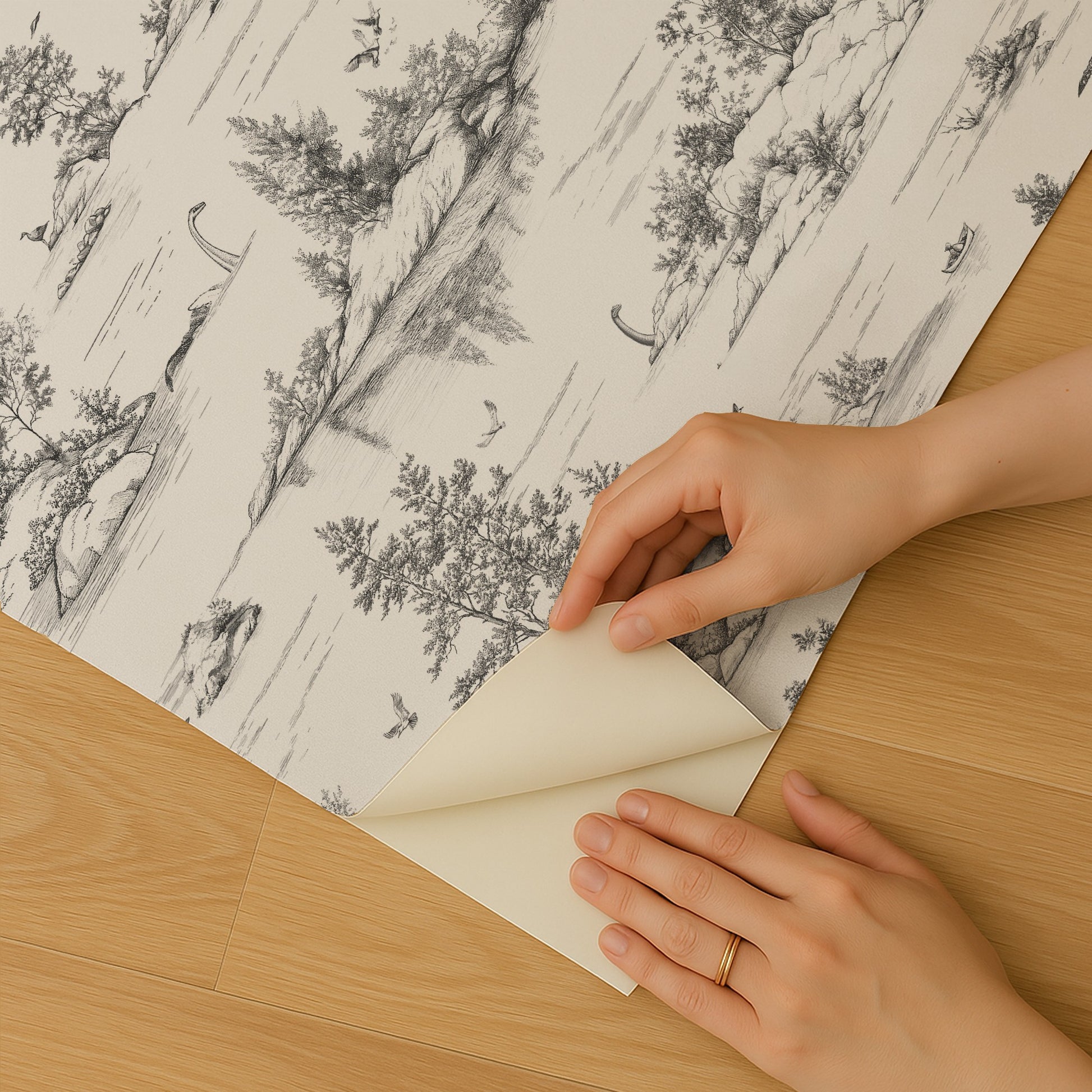 Grayscale forest wallpaper sheet being peeled back to reveal backing, hands with a ring on a wooden floor.