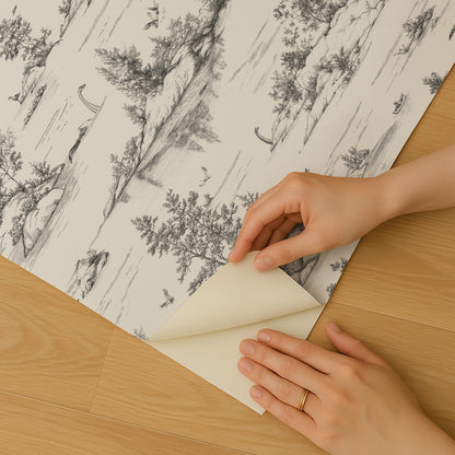 Grayscale forest wallpaper sheet being peeled back to reveal backing, hands with a ring on a wooden floor.