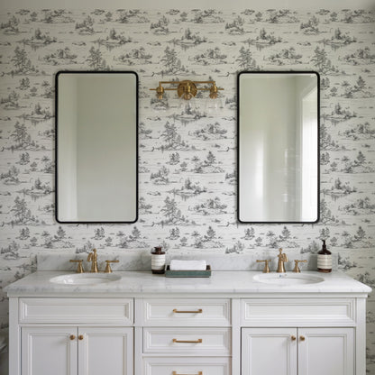White double-sink bathroom vanity with marble countertop, brass hardware, black-framed mirrors, and toile wallpaper.