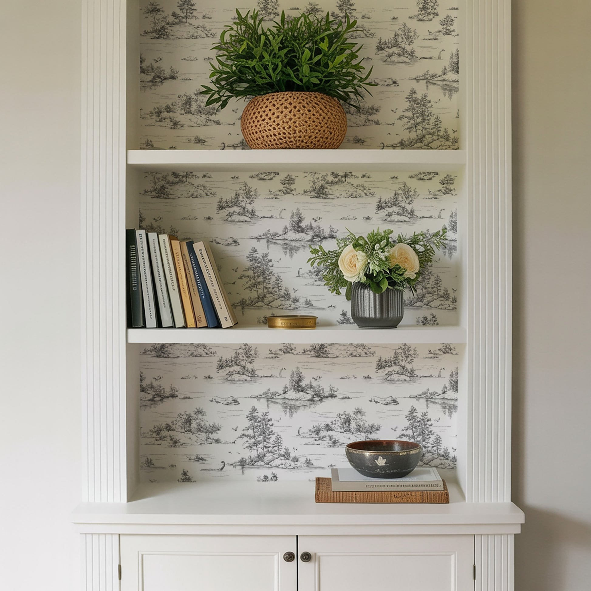 White built-in bookshelf with greenery in a woven basket, books, a vase of flowers, and a bowl on a stack of books.