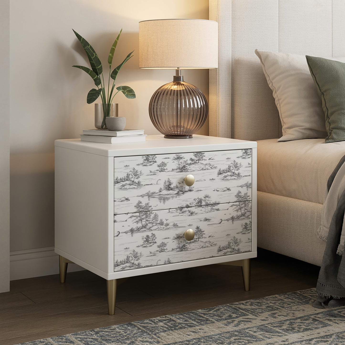 White nightstand with two drawers in grey toile pattern, brass knobs, tapered legs; lamp, plant & books on top.