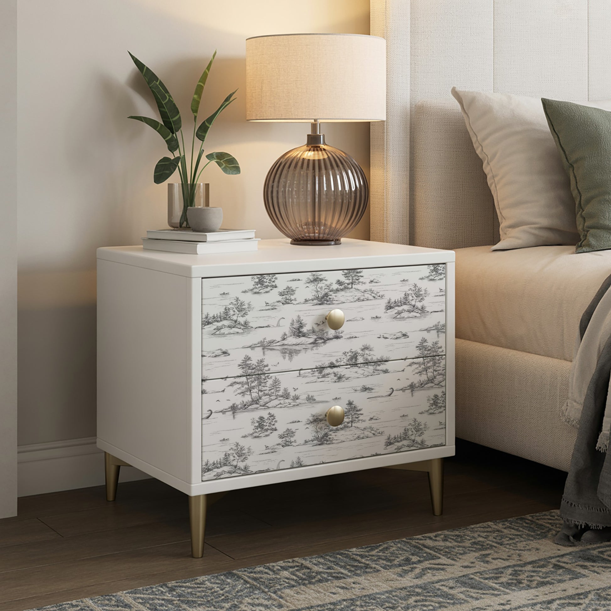 White nightstand with two drawers in grey toile pattern, brass knobs, tapered legs; lamp, plant & books on top.
