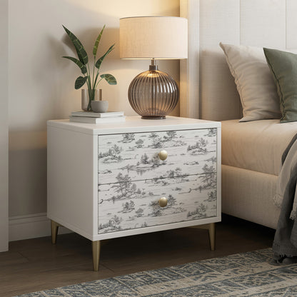 White nightstand with two drawers in grey toile pattern, brass knobs, tapered legs; lamp, plant & books on top.