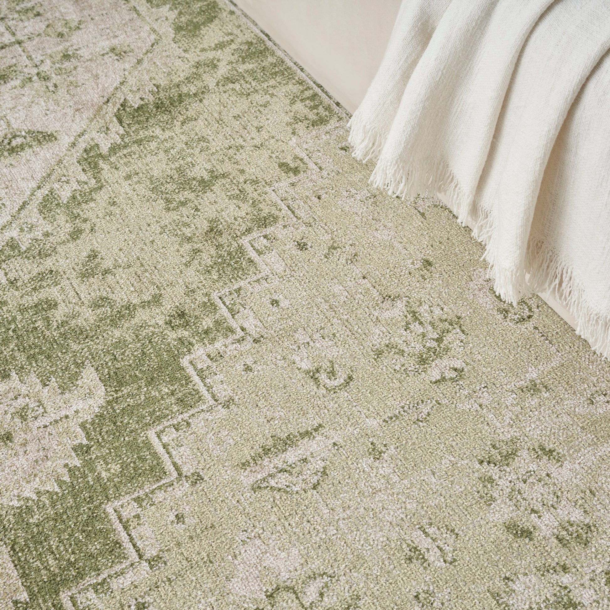 Olive green and beige distressed oriental rug with geometric border; woven texture, cream fringe throw draped at edge.
