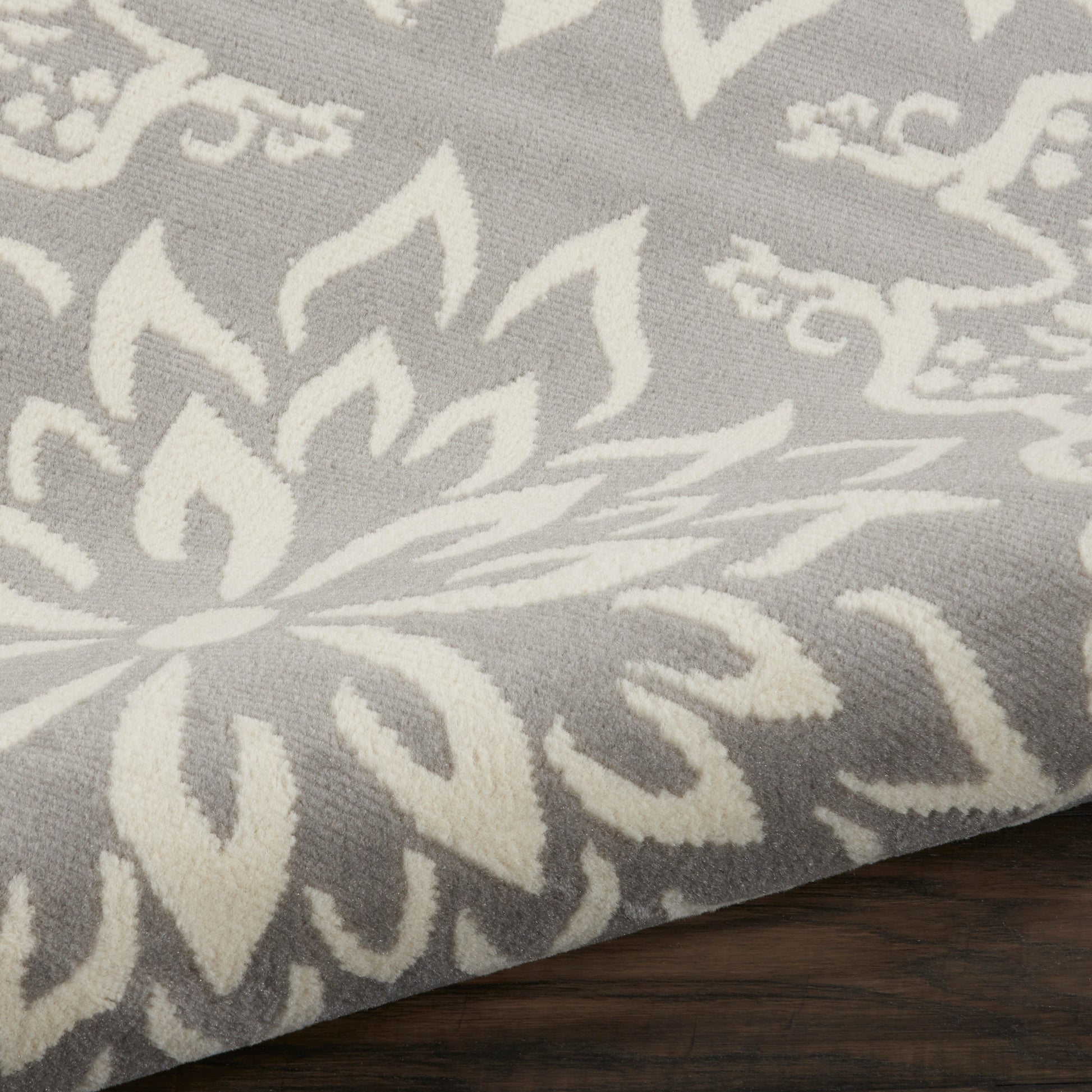 Gray rug with white damask floral pattern; soft woven texture and a curled corner on dark wood floor.