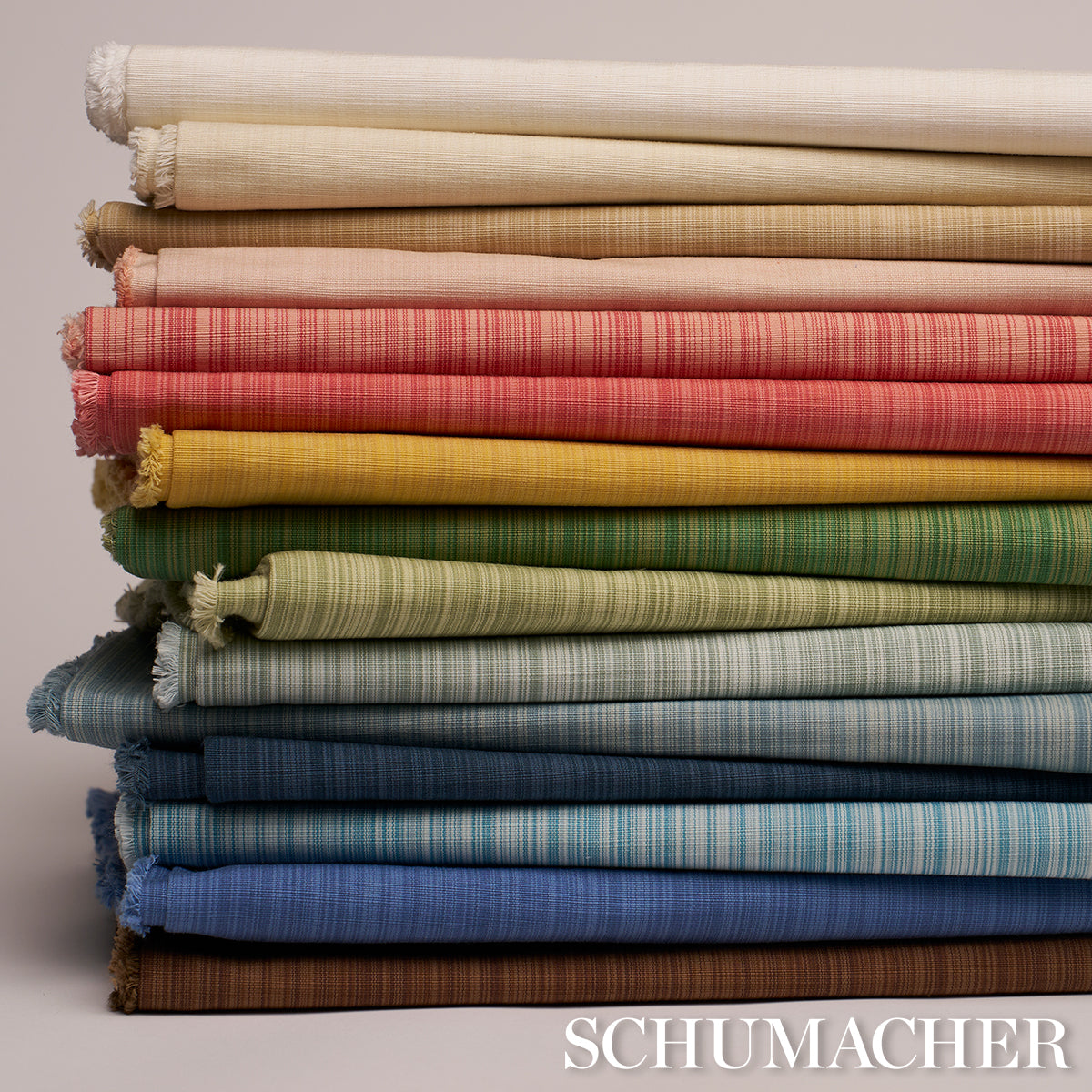 Stack of rolled woven fabric bolts in a rainbow of colors, fringed edges, linen/cotton texture, Schumacher label visible.