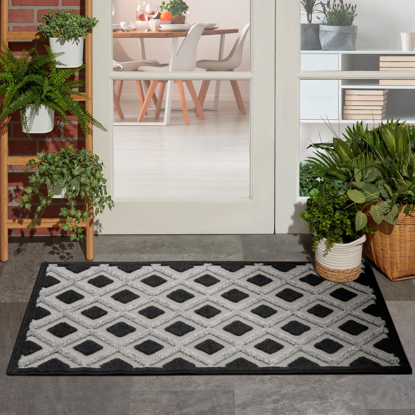 Entry rug with white diamond lattice on black background; plants nearby and a modern dining room visible through glass doors.