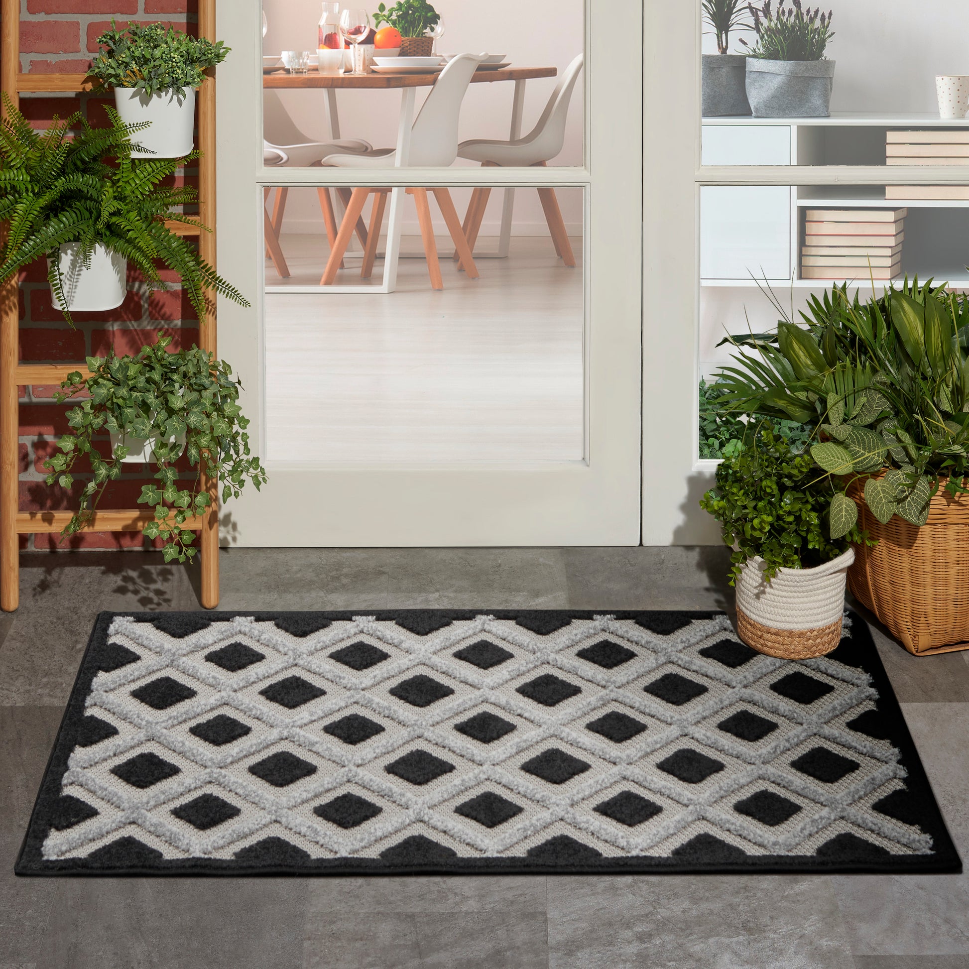 Entry rug with white diamond lattice on black background; plants nearby and a modern dining room visible through glass doors.