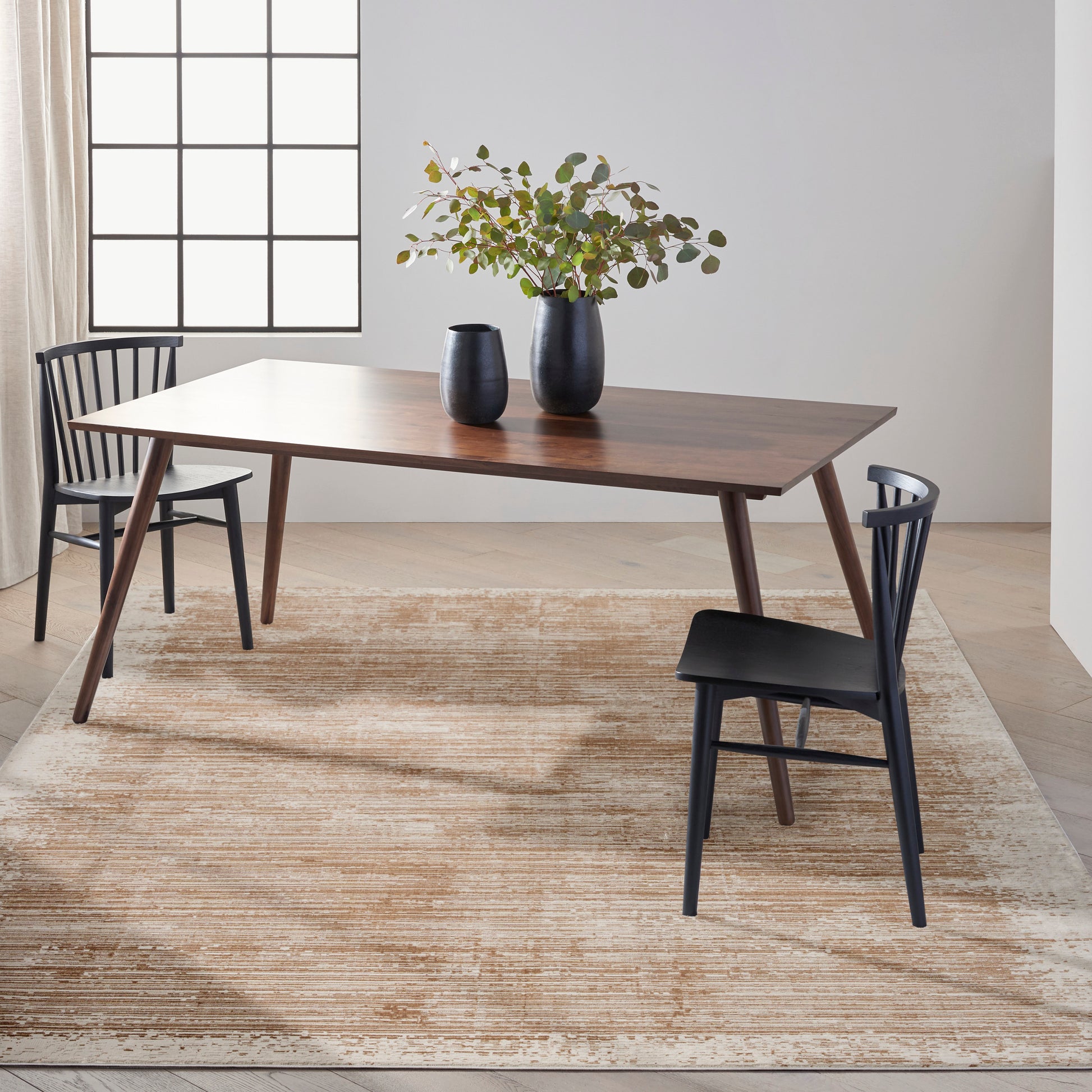 Mid-century wooden dining table with dark finish & black chairs, black vases with greenery on beige rug.