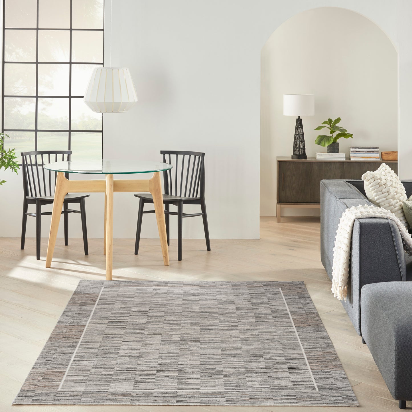 Grey-bordered rug (6' x 9') in a bright living room with a round glass dining table, natural wood legs, and two black chairs.