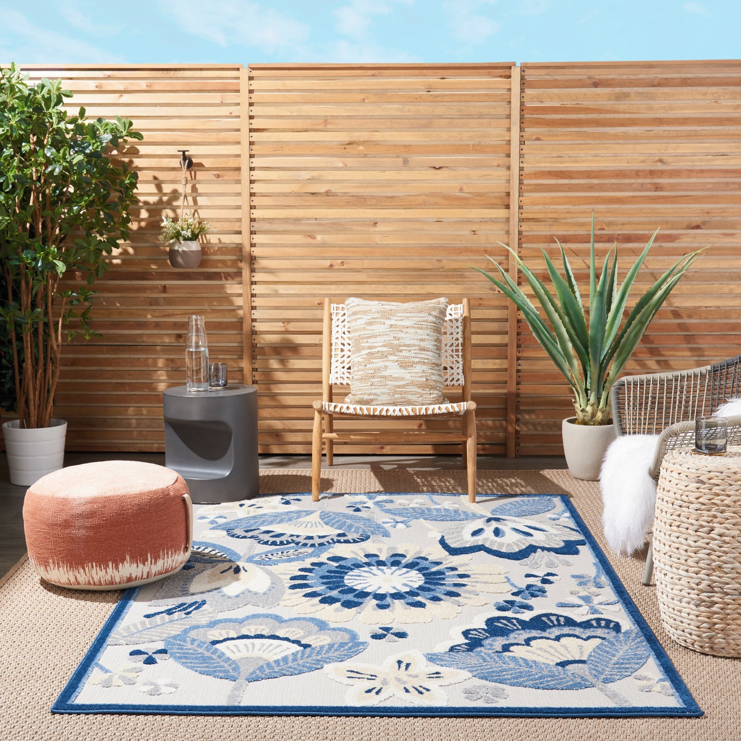 Outdoor patio with blue floral rug, wicker chair, round peach pouf, potted plants, and a wooden slat fence.