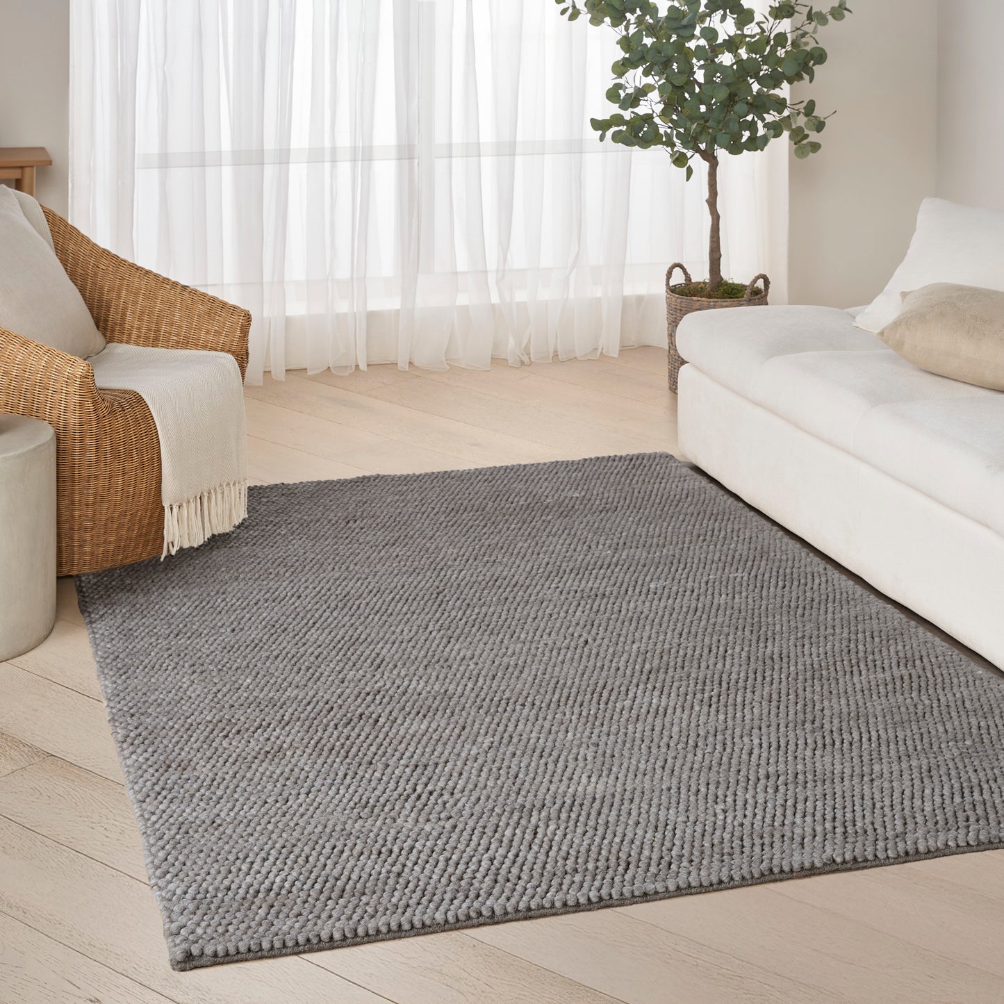 Rectangular light-gray woven rug in a bright living room with a white sofa, wicker chair, sheer curtains, and a potted plant.
