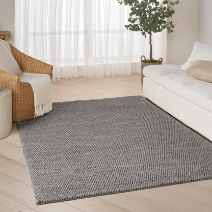 Rectangular light-gray woven rug in a bright living room with a white sofa, wicker chair, sheer curtains, and a potted plant.