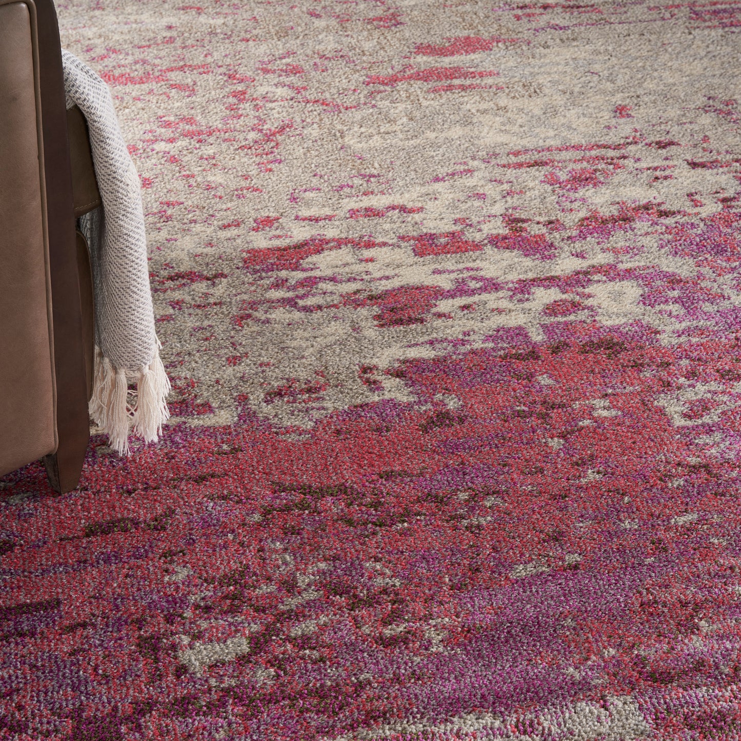 Abstract pink and purple area rug with beige tones, beside a brown leather sofa and cream knit throw.