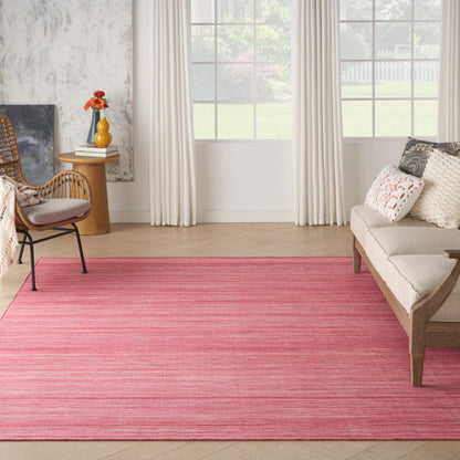 Living room scene with a bright pink rug, beige sofa, wicker chair, round wooden side table with orange flowers, and a large window with white curtains.
