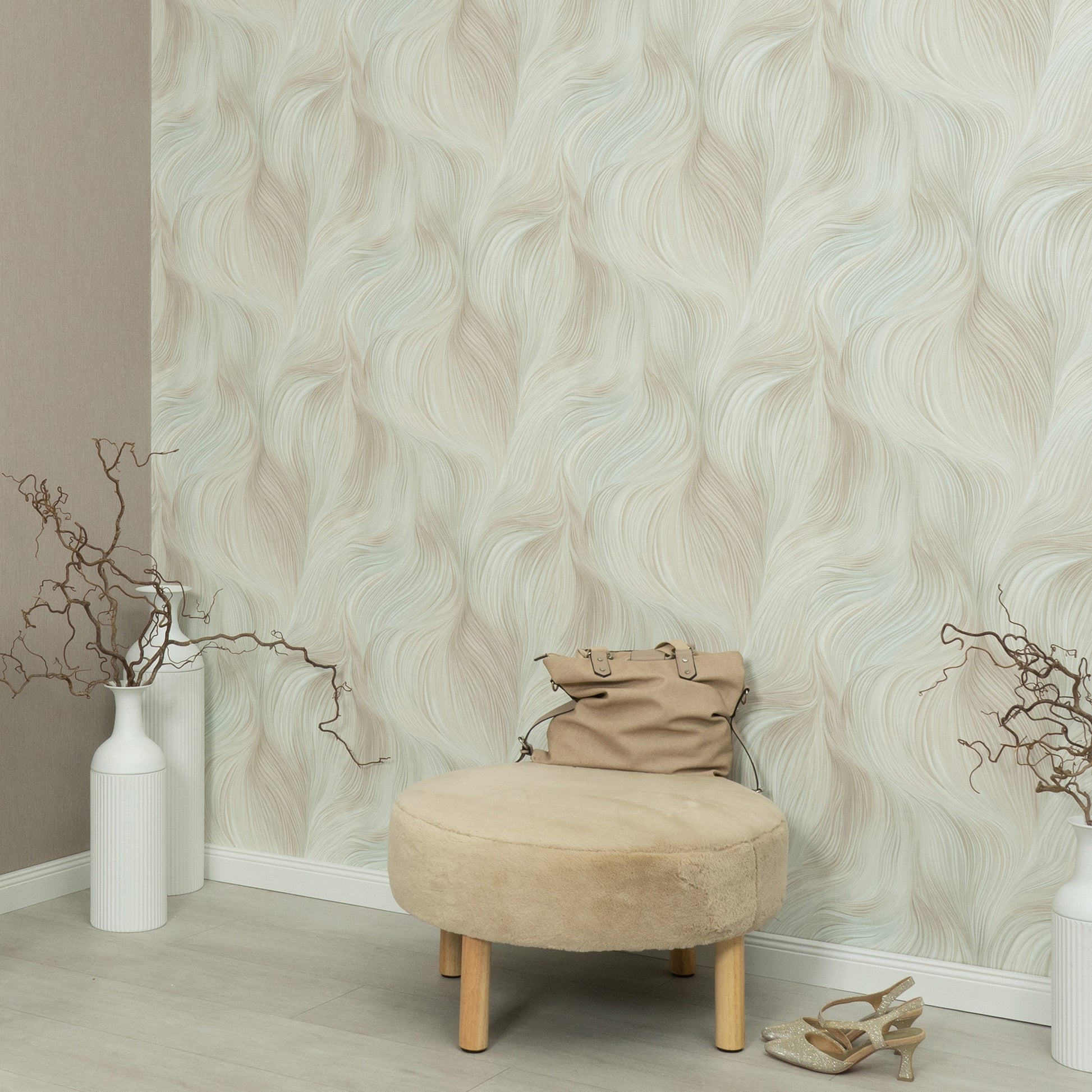 Teal wallpaper with a soft, wavy light pattern on the wall; foreground shows a beige round ottoman with wooden legs, a tan handbag on top, white vases with bare branches, and sandals nearby.