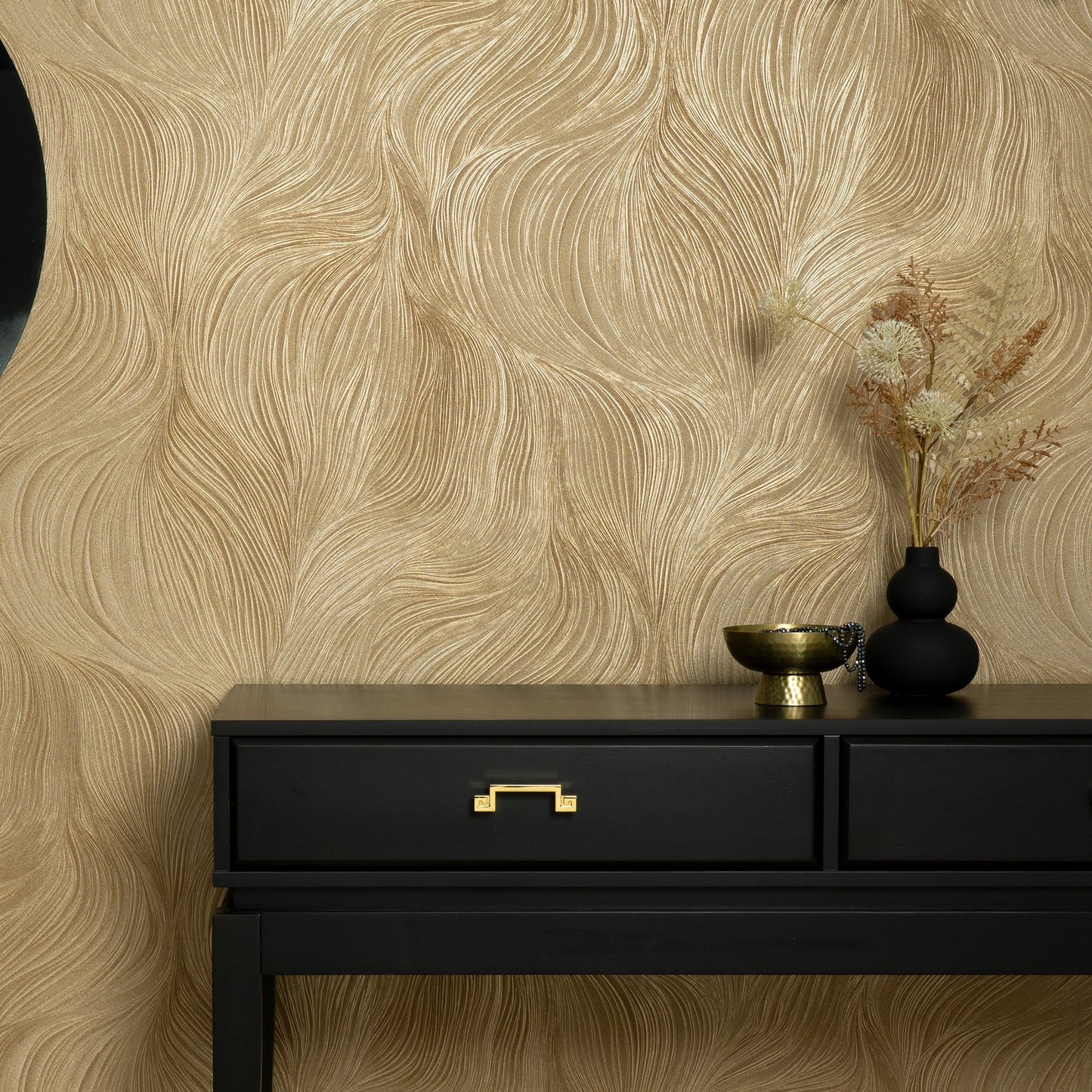 Black two-drawer console with gold handle, gold bowl, black vase and dried stems against gold textured wallpaper.