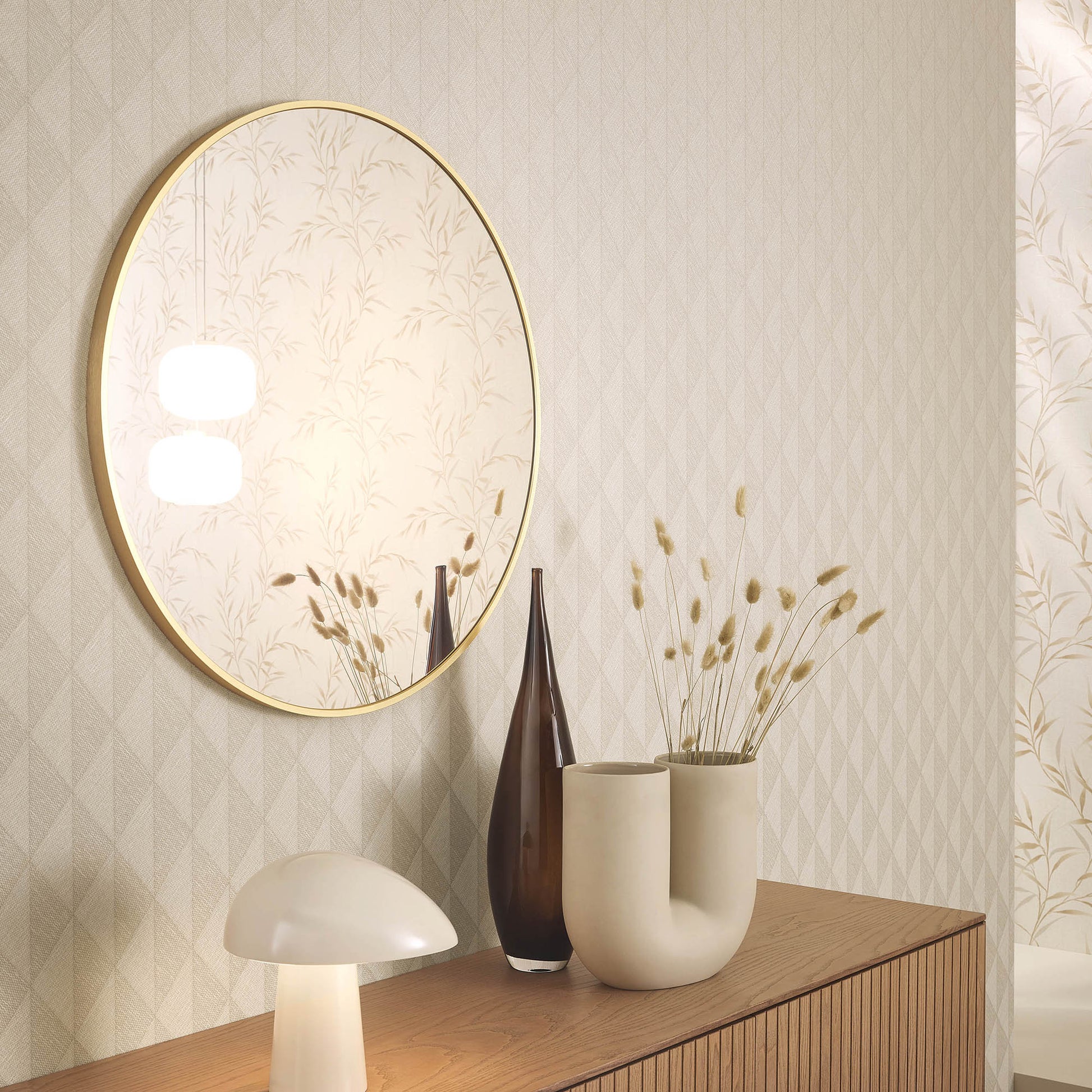 Round gold-framed mirror above a wooden sideboard with a mushroom lamp, brown glass vase and cream ceramic vases with dried grasses.