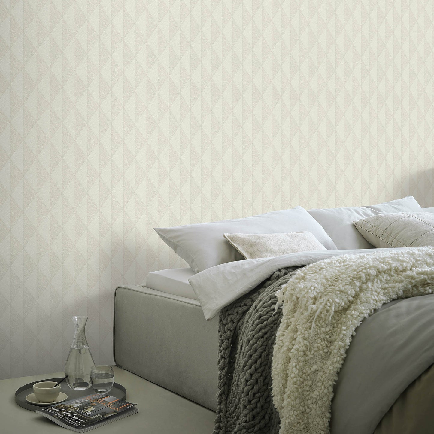 Neutral bedroom with taupe wallpaper on the walls, a gray upholstered bed, cream knit throw, white pillows, and a glass carafe & cup on a tray.