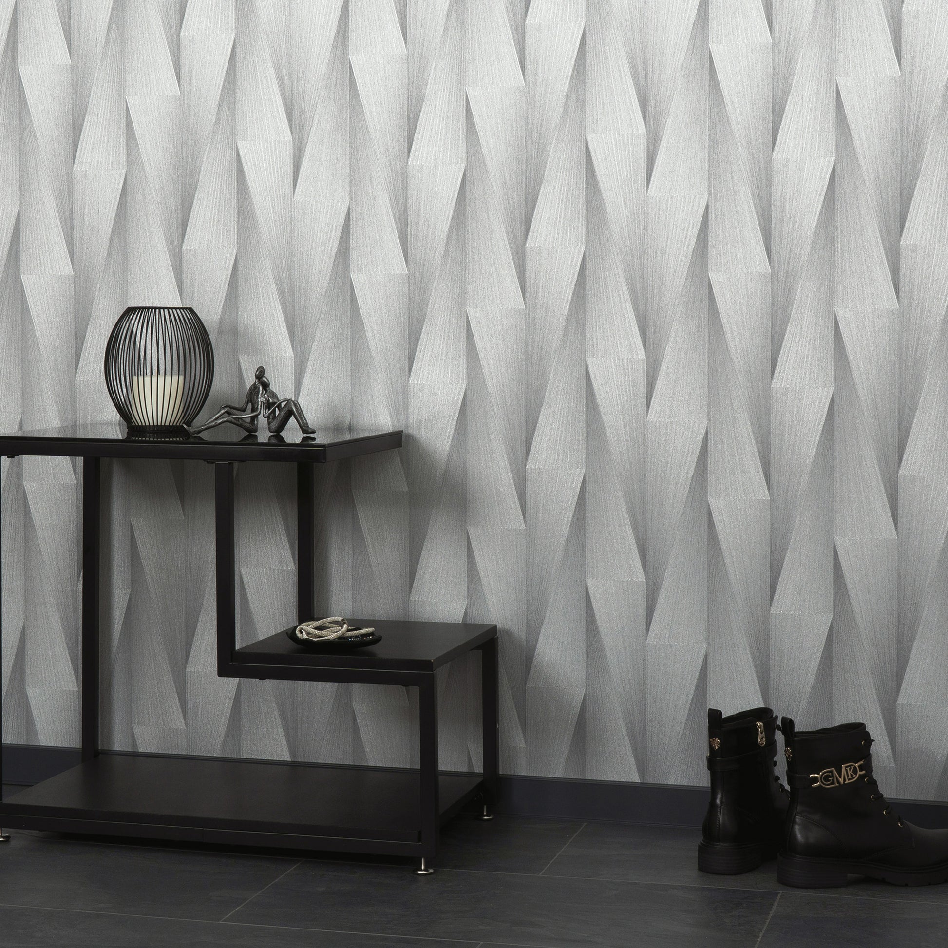 Geometric gray wallpaper behind a black stepped console table with a lantern lamp, small figurine, rope coil, and black buckle ankle boots on dark tile.