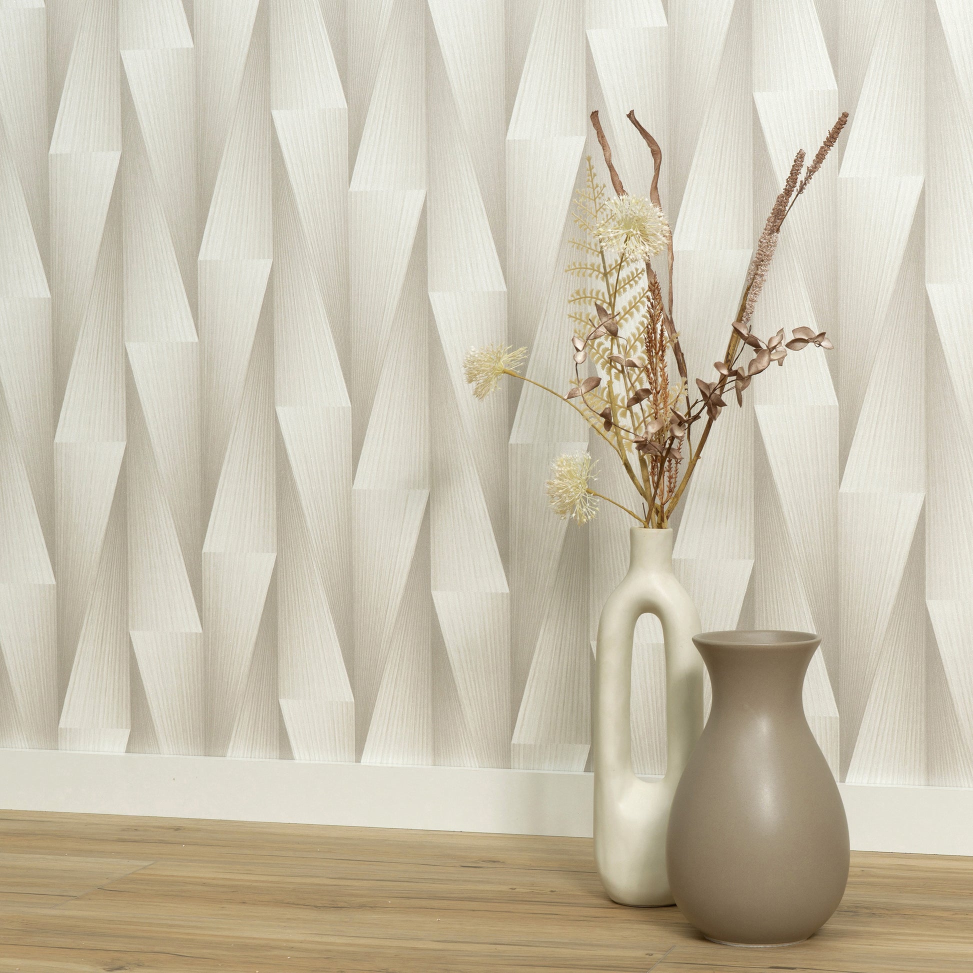 Tall cream vase with handle and dried grasses beside a taupe rounded vase on a light wood floor against geometric wallpaper.