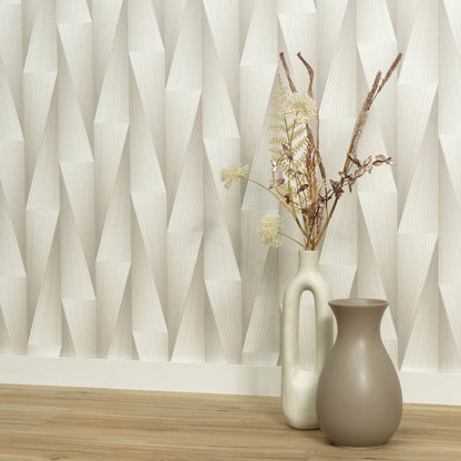 Tall cream vase with handle and dried grasses beside a taupe rounded vase on a light wood floor against geometric wallpaper.