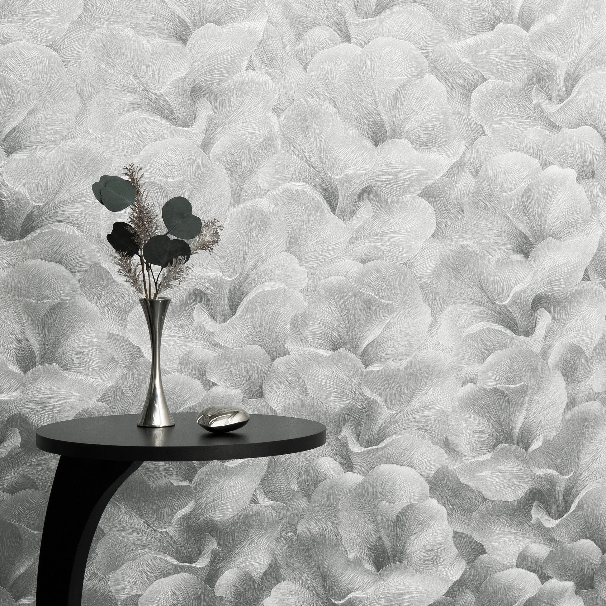Black round side table with curved leg, silver vase of dried greenery & a metallic dish against grey botanical wallpaper.