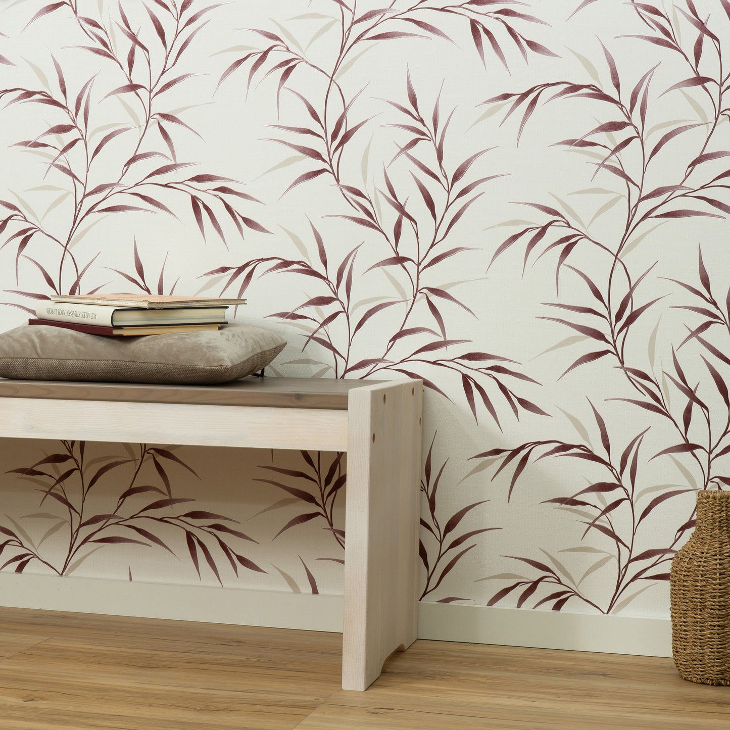 Light wooden bench with a beige cushion and stacked books, against cream wallpaper with burgundy botanical leaves; woven basket nearby.