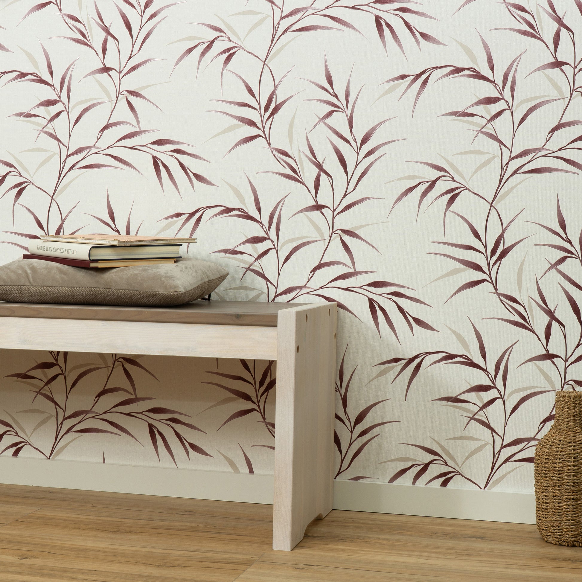 Light wooden bench with a beige cushion and stacked books, against cream wallpaper with burgundy botanical leaves; woven basket nearby.