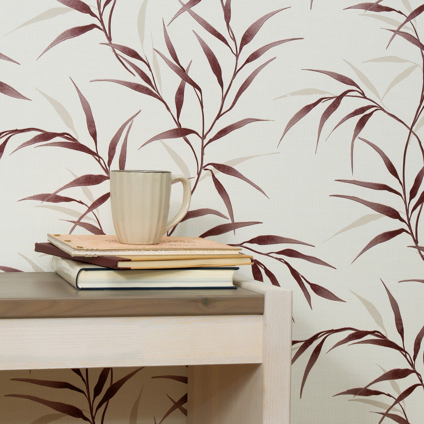 Beige ceramic mug on a stack of notebooks atop a light wood desk against burgundy leaf-pattern wallpaper.
