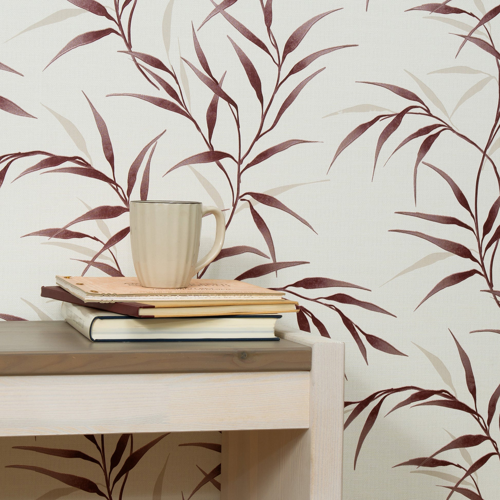 Beige ceramic mug on a stack of notebooks atop a light wood desk against burgundy leaf-pattern wallpaper.