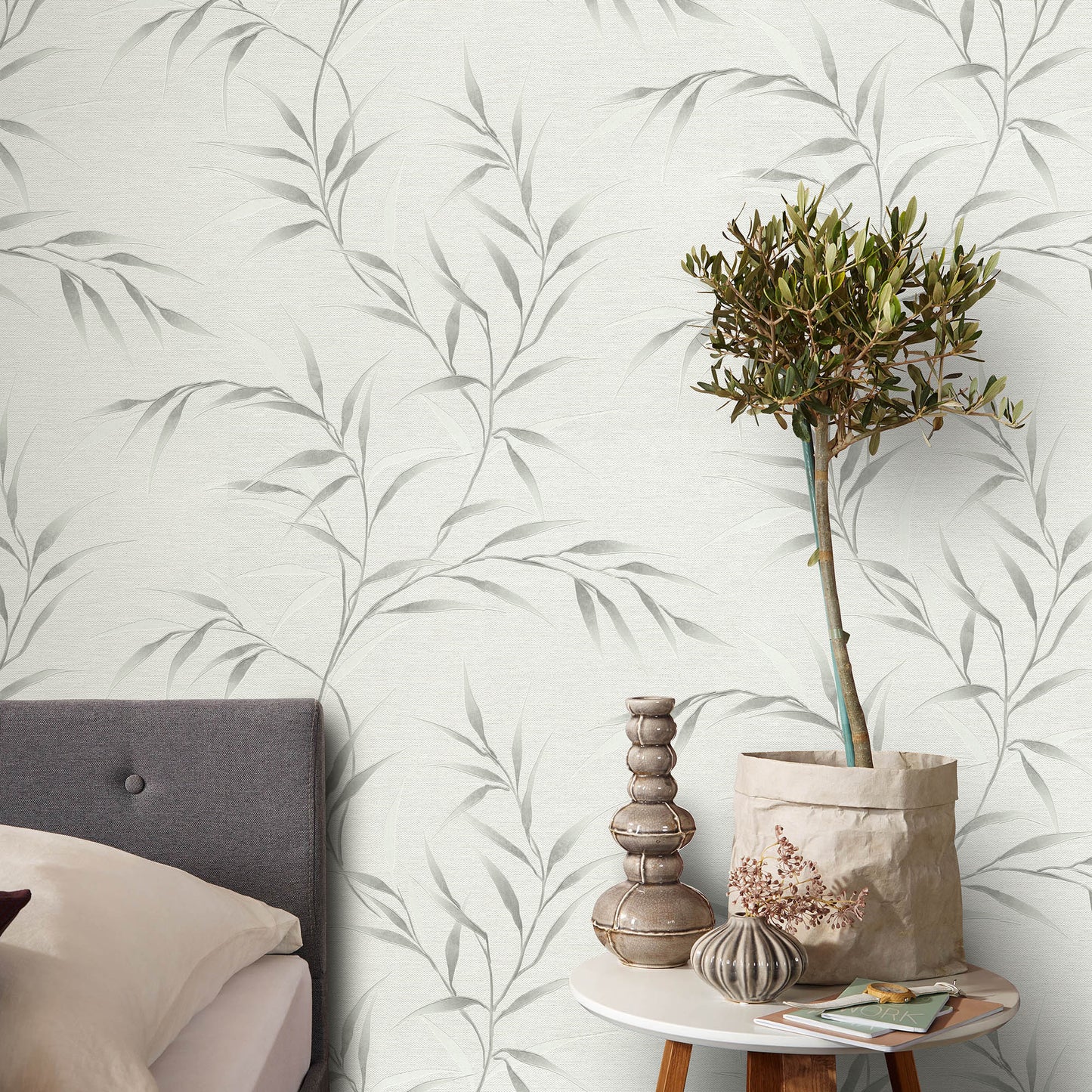 Cozy bedroom with pale botanical wallpaper, gray upholstered headboard, and round table with a potted tree and vases.