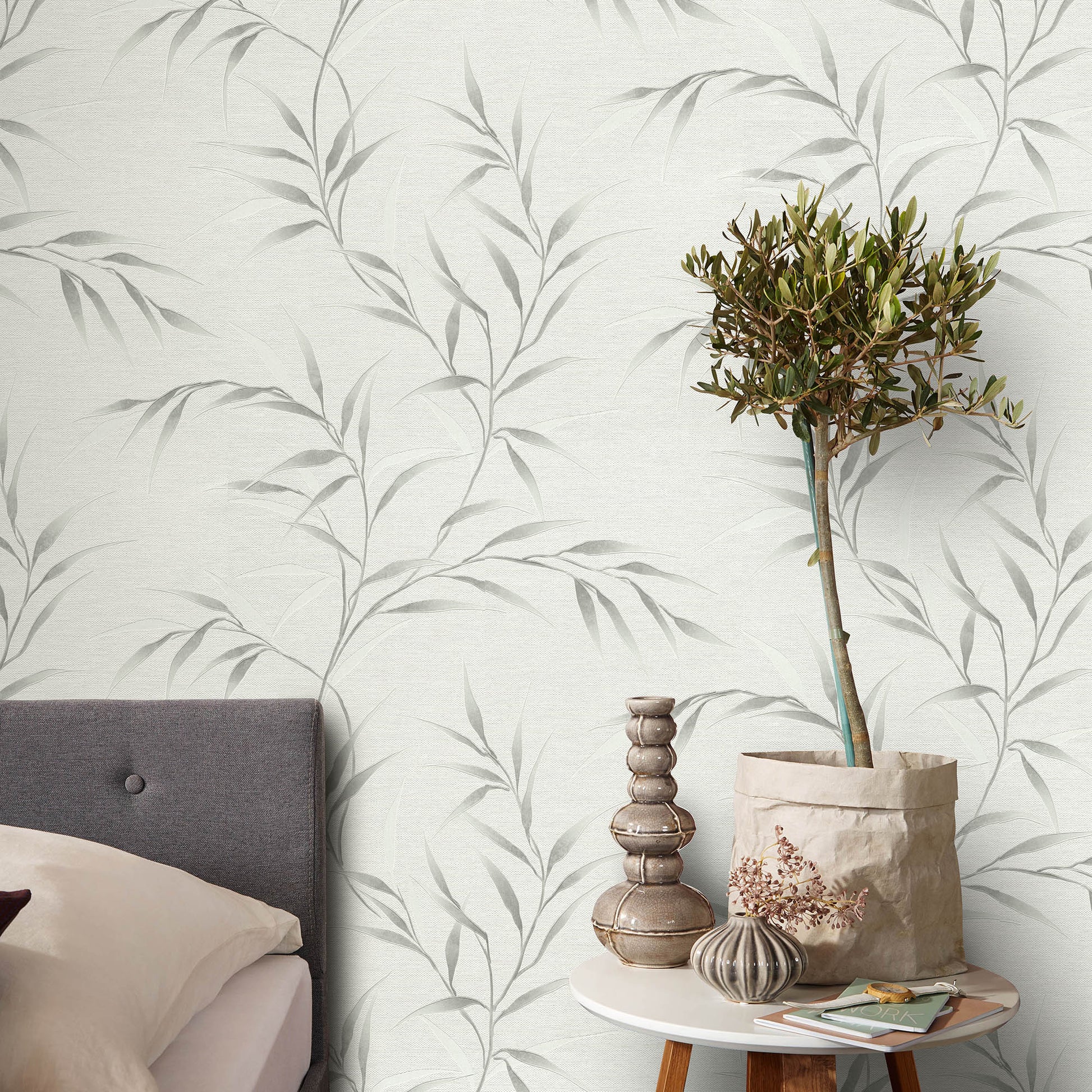 Cozy bedroom with pale botanical wallpaper, gray upholstered headboard, and round table with a potted tree and vases.