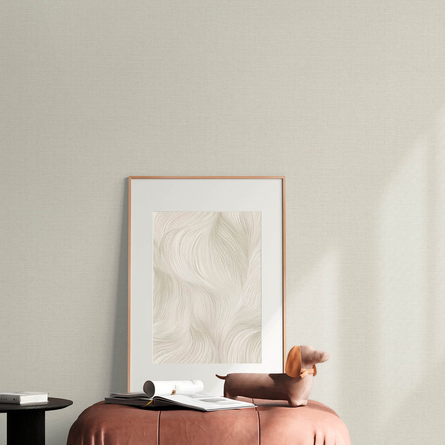 Beige wall with sepia jute texture wallpaper featuring a subtle abstract pattern; foreground shows a brown leather ottoman, a small brown sculpture & an open book.