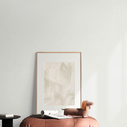 Beige framed abstract art leaning on pale wall; soft pink ottoman beside round black side table with books.