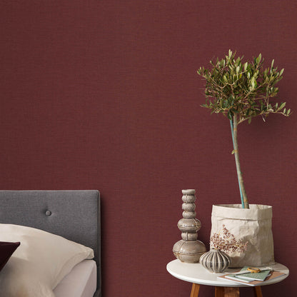 Burgundy red jute-textured wallpaper on bedroom walls, with a gray headboard and a round table displaying an olive tree in a paper bag, a wooden bead sculpture, a vase, and magazines.