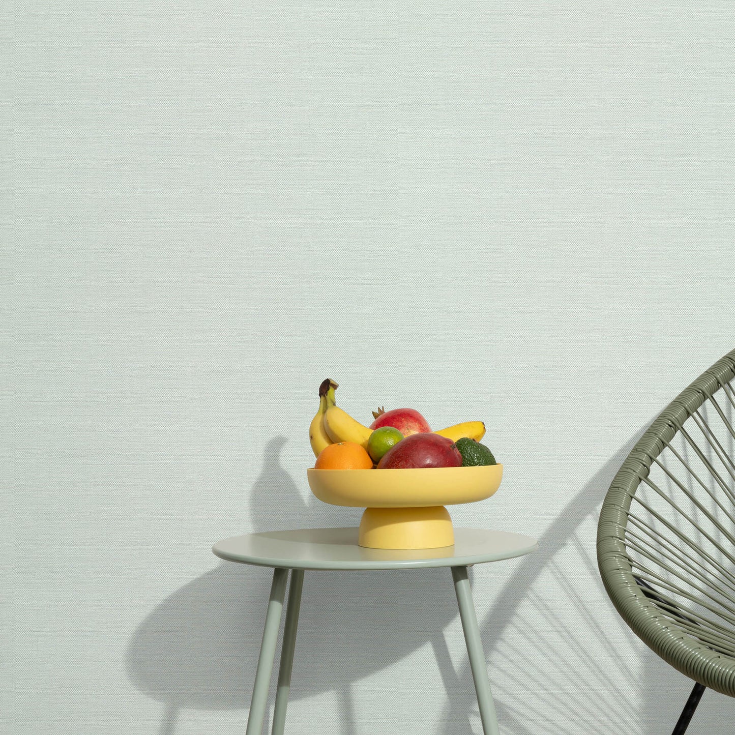 Yellow pedestal fruit bowl on a white round table with bananas, apples and citrus; minimalist scene with a woven chair nearby