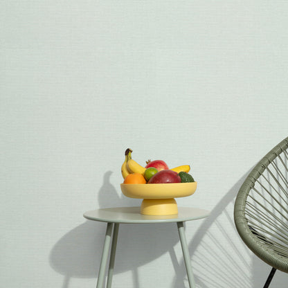 Yellow pedestal fruit bowl on a white round table with bananas, apples and citrus; minimalist scene with a woven chair nearby