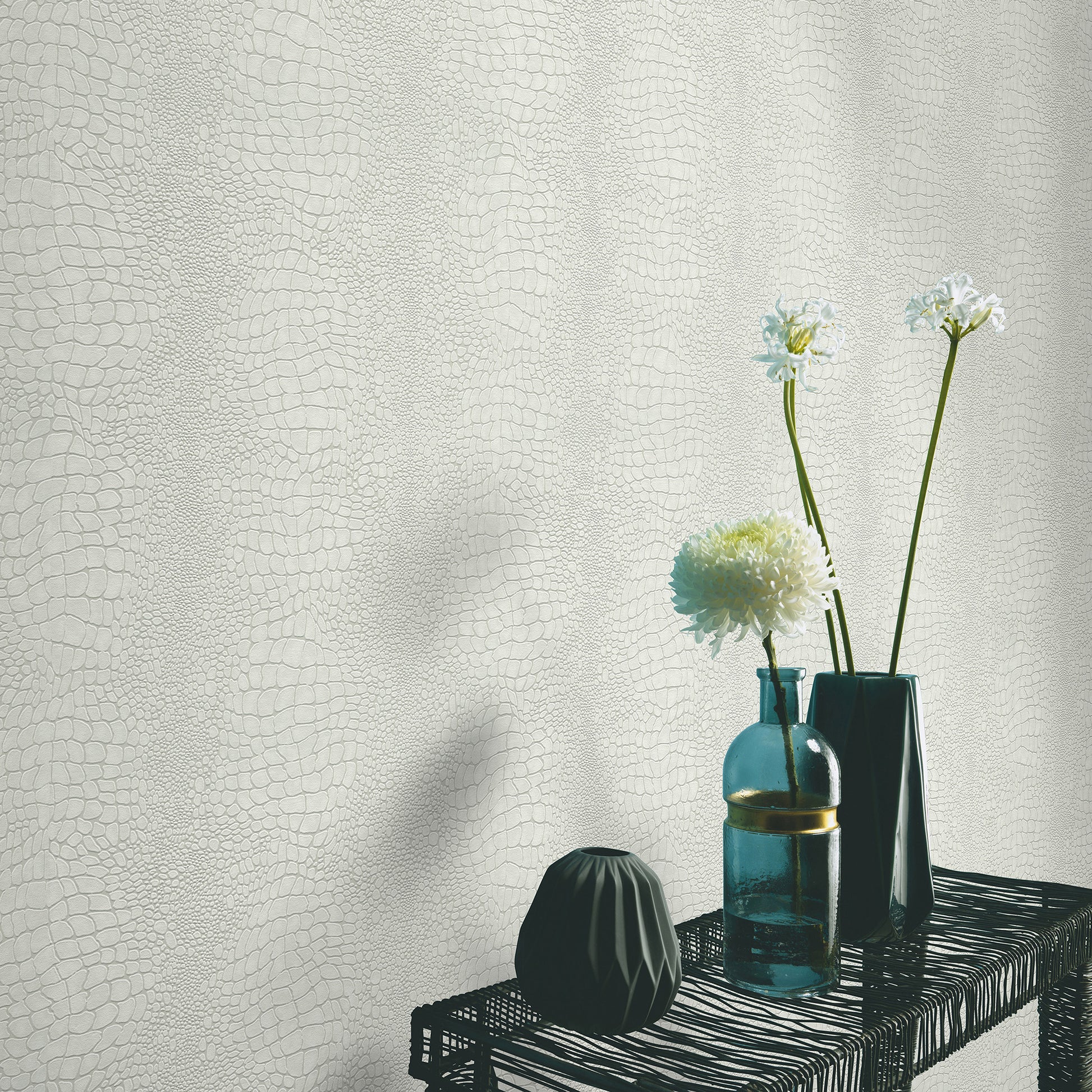 Beige textured backdrop with a black woven shelf holding two teal glass bottles, a faceted black vase, and white blooms.
