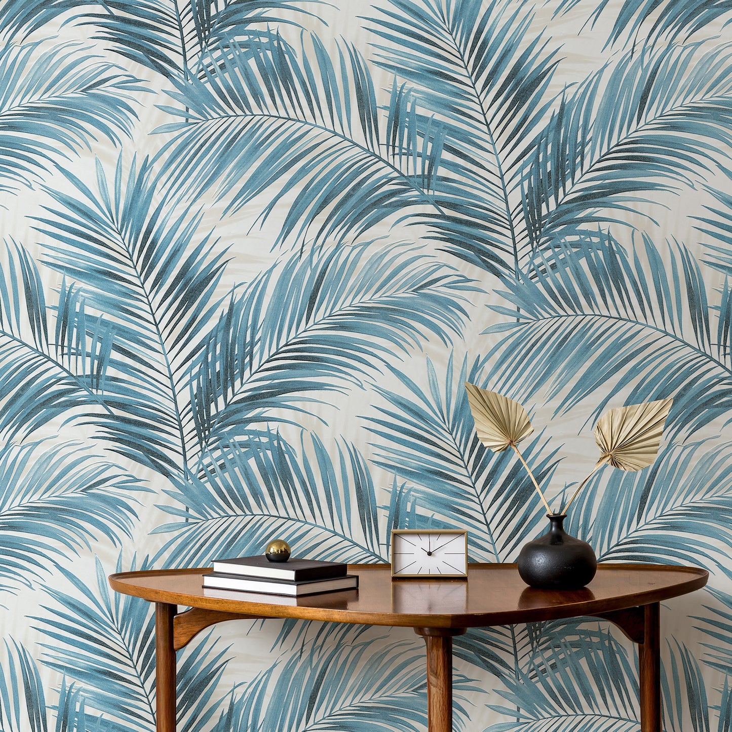 Mid-century oval wooden console table beside blue palm-leaf wallpaper; books, clock, and black vase with golden palm fronds.